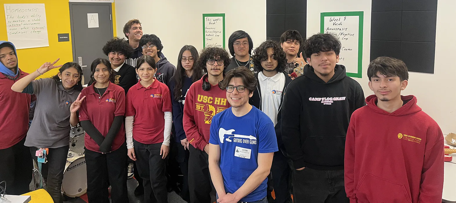 Students and a mentor from Guitars Over Guns posing together in a classroom at USC Hybrid High School, wearing school shirts and hoodies.
